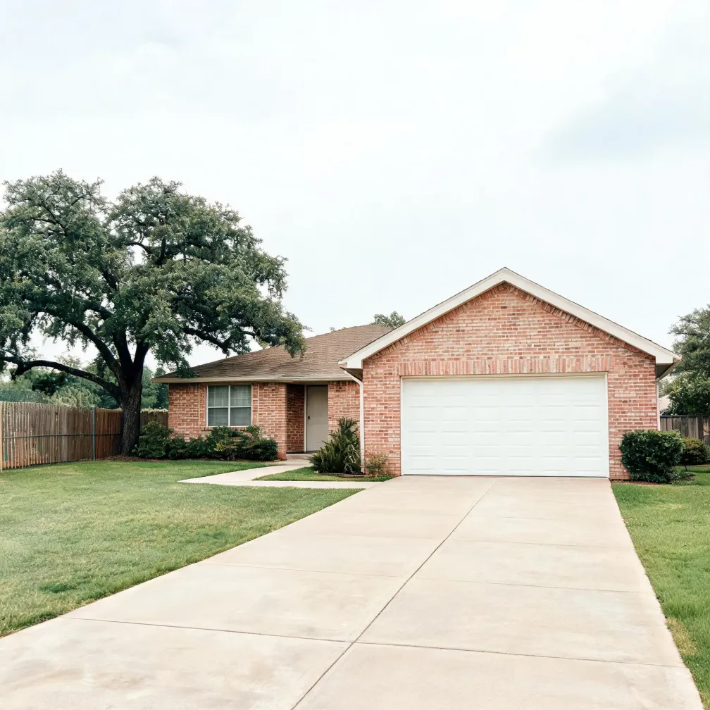 Single-story red brick house with a white two-car garage door, a wide concrete driveway leading to it, a mown lawn, a wooden fence at left, and a large oak tree beside the house under an overcast sky.