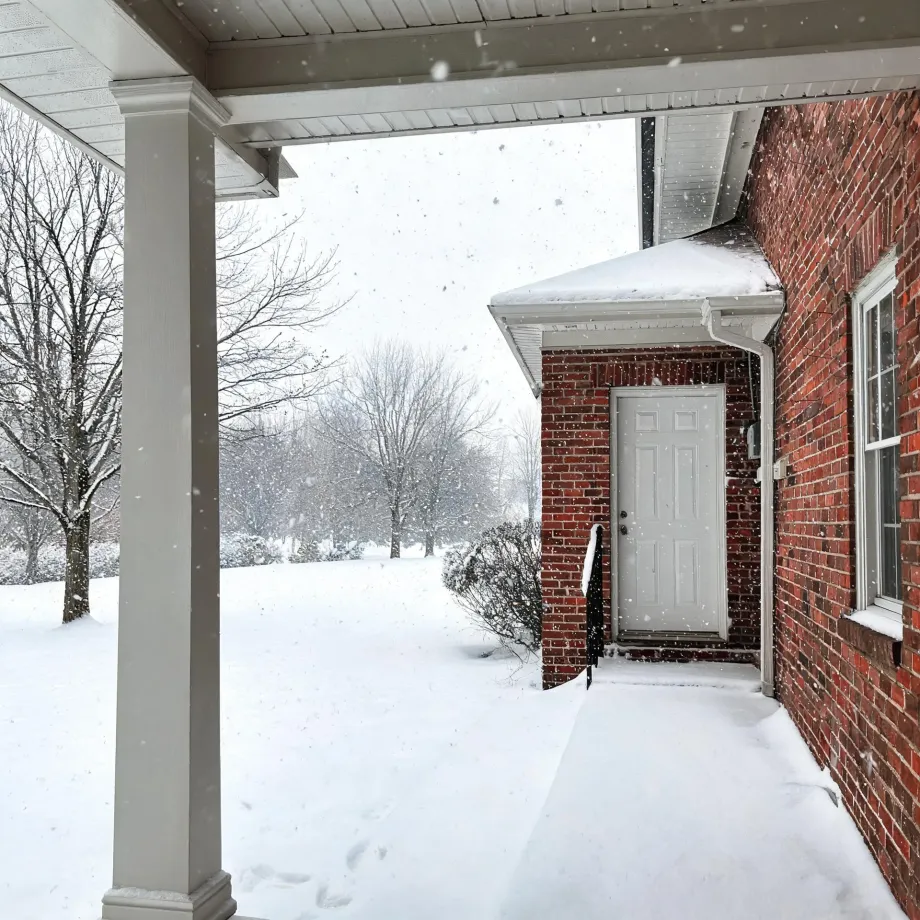View from a covered porch with a white column toward a red brick house wall with a white side door and window, during falling snow over a snow-covered yard with bare trees.