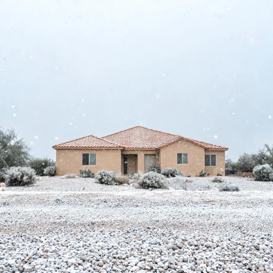 A single-story tan stucco house with a red tile roof stands behind snow-dusted shrubs and a gravel yard under a pale sky with falling snow.