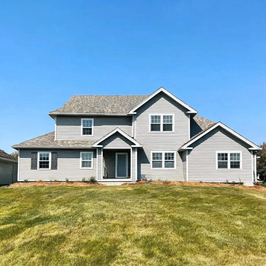 A two-story house with gray and white horizontal siding, multiple gables, asphalt shingle roof and a small covered front entry, fronted by a large green lawn under a clear blue sky.
