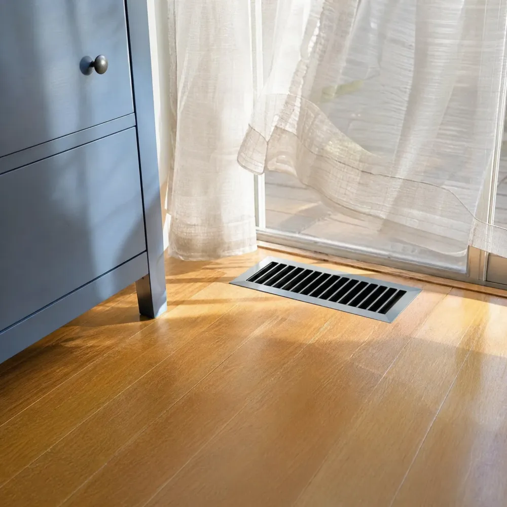 A metal louvered floor register set into a light wood plank floor near a glass door with sheer curtains, beside a gray cabinet with a round knob.