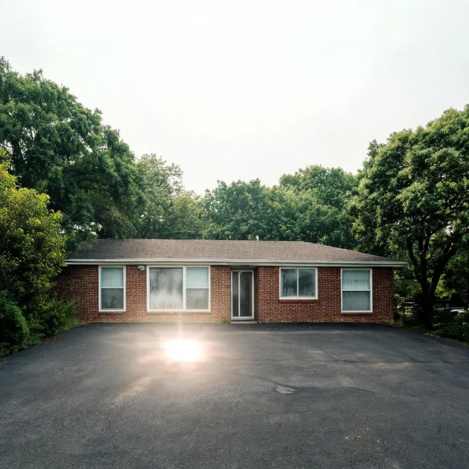 A single-story red brick ranch house with a low shingled roof, white-framed windows and a central glass door, fronted by a large empty asphalt parking area with sun glare, surrounded by green deciduous trees under a pale overcast sky.