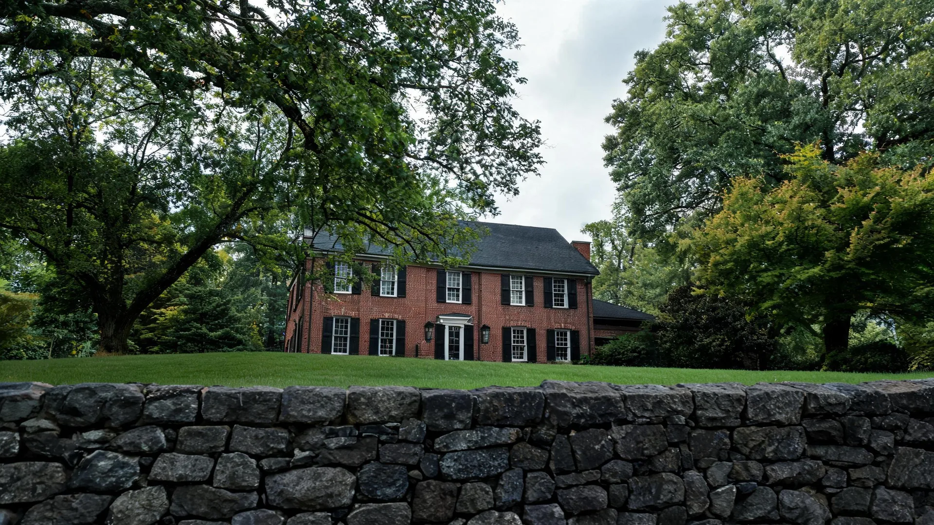 A two-story red brick house with black shutters and a dark roof sits behind a mown lawn, large deciduous trees, and a dry-stacked stone wall in the foreground.