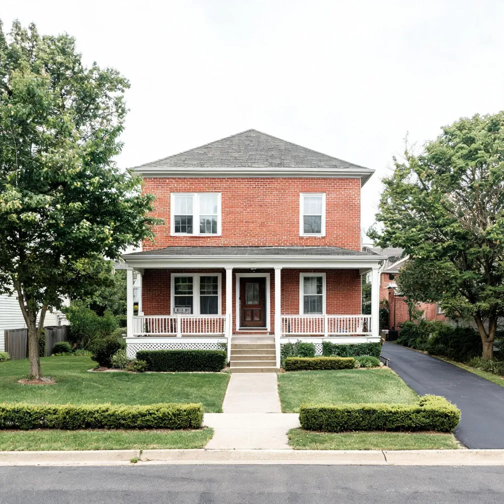A two-story red brick house with a white covered front porch, gray hipped roof, white-framed windows, a concrete walkway, trimmed hedges, lawn, and an asphalt driveway at right, flanked by deciduous trees under an overcast sky.