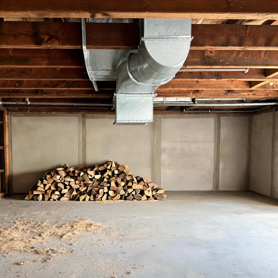 An unfinished basement with exposed wood floor joists, galvanized sheet-metal HVAC ductwork and pipes overhead, smooth gray concrete walls, and a stack of split firewood on the concrete floor with sawdust scattered nearby.