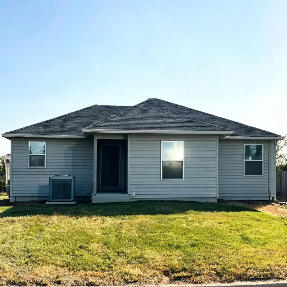 Single-story house with light gray horizontal siding, dark shingled hip roof, a dark front door with a small concrete stoop, three windows, and an air conditioning condenser unit at left, fronted by a grass lawn under a clear sky.