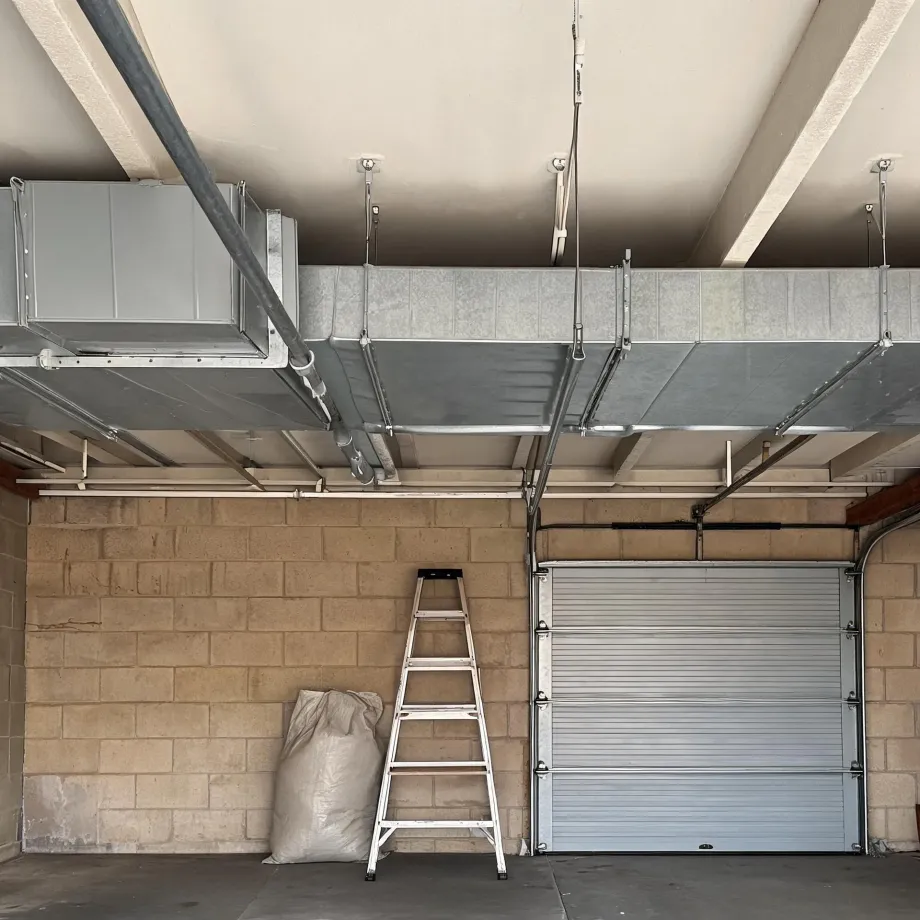 Interior of a concrete block garage with exposed galvanized sheet metal ductwork suspended from the ceiling, a white metal stepladder and a filled bag leaning against the wall, and a closed sectional roll-up door on the right.