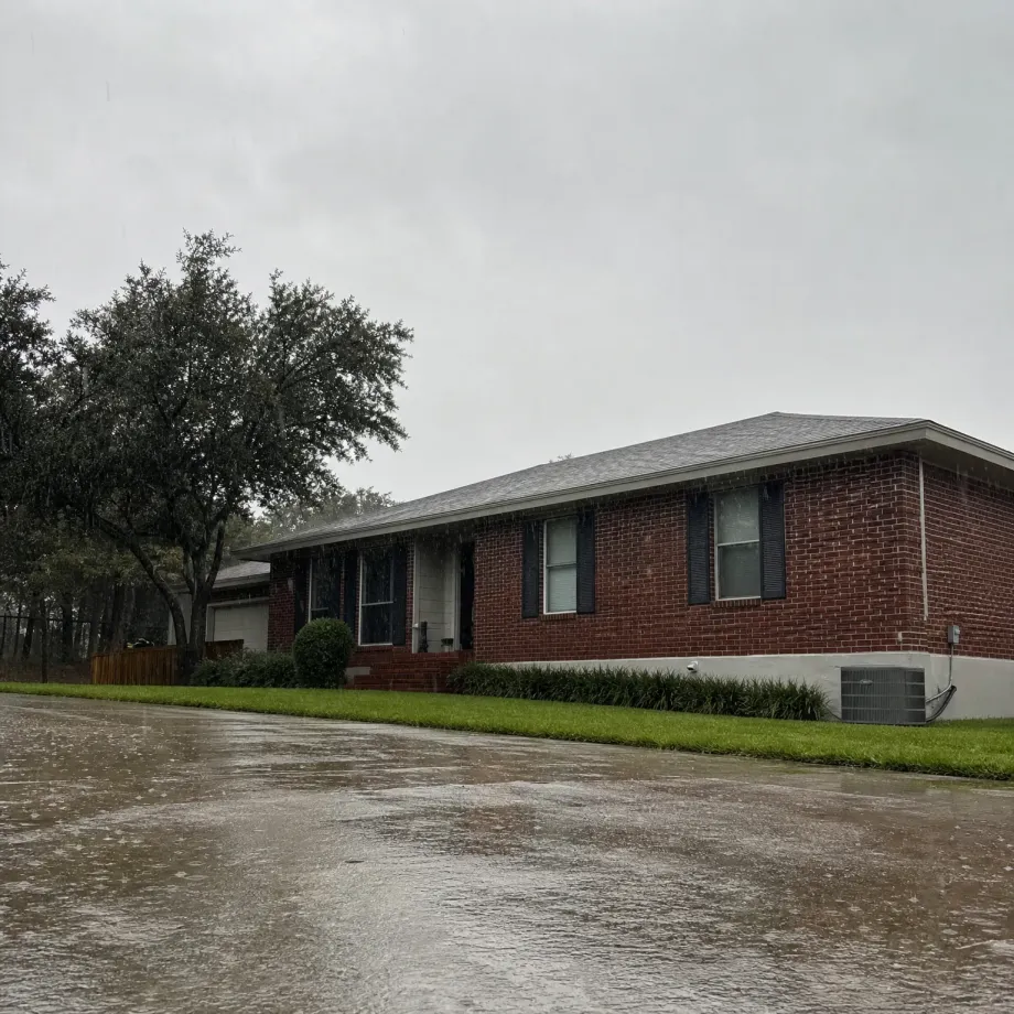 A single-story red brick house with a gray shingled roof and dark shutters during heavy rain, with water streaming off the eaves, a wet street in the foreground, green lawn and shrubs, and large oak trees to the left under an overcast sky.