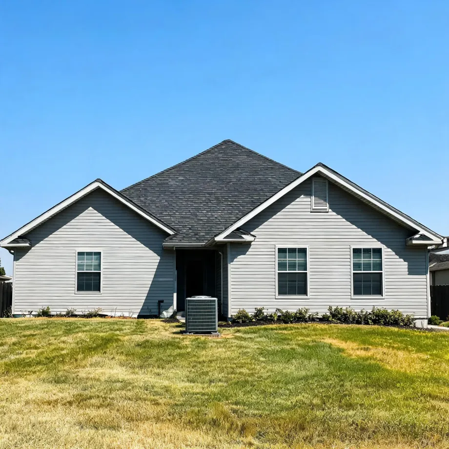 A single-story house with light gray horizontal siding, dark shingled gable roofs, and white-trimmed windows, with an air conditioning unit beside a recessed entry and a partly dry lawn in front under a clear blue sky.