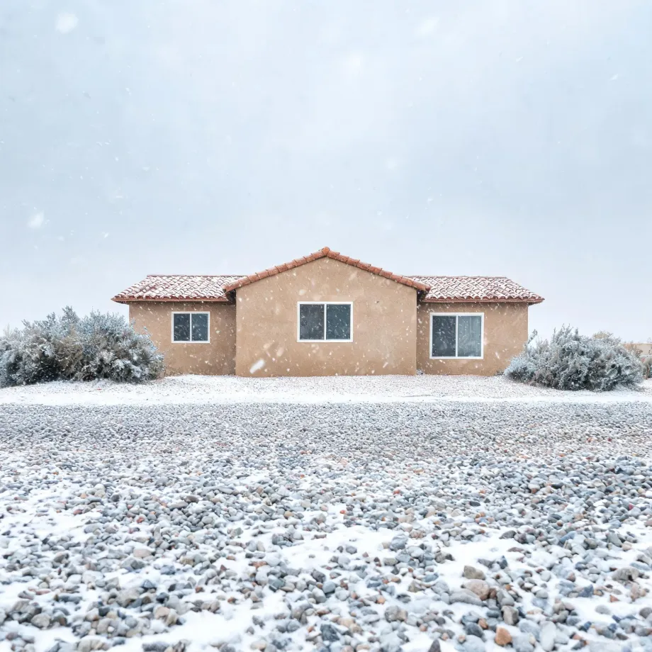 A single-story tan stucco house with a red tile roof and three windows, flanked by frost-covered shrubs, behind a wide gravel yard dusted with snow under a pale overcast sky with falling snowflakes.