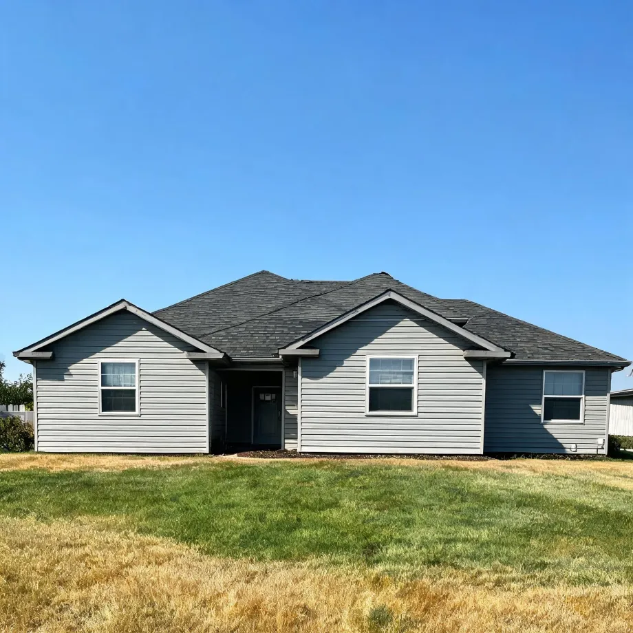 Single-story house with gray horizontal lap siding, dark asphalt shingle roof, two front-facing gables, a recessed entry door and white-trimmed windows, fronted by a patchy green and brown lawn under a clear blue sky.