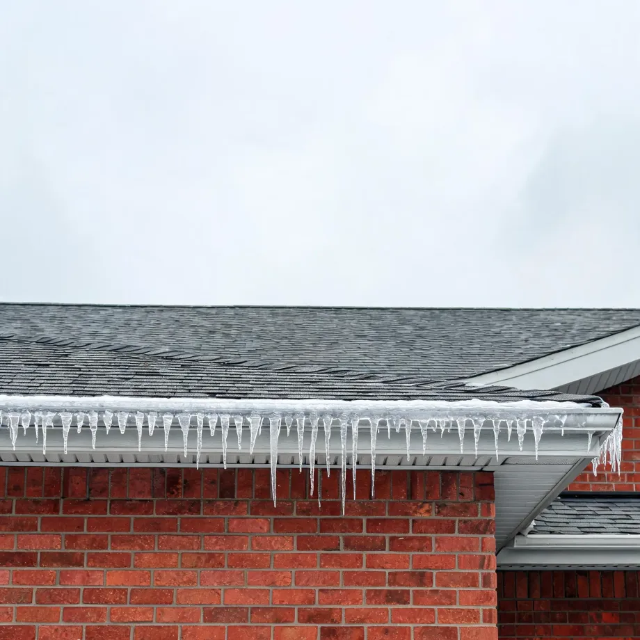 Icicles hanging from a white gutter along the edge of a gray shingled roof on a red brick house, under an overcast sky.