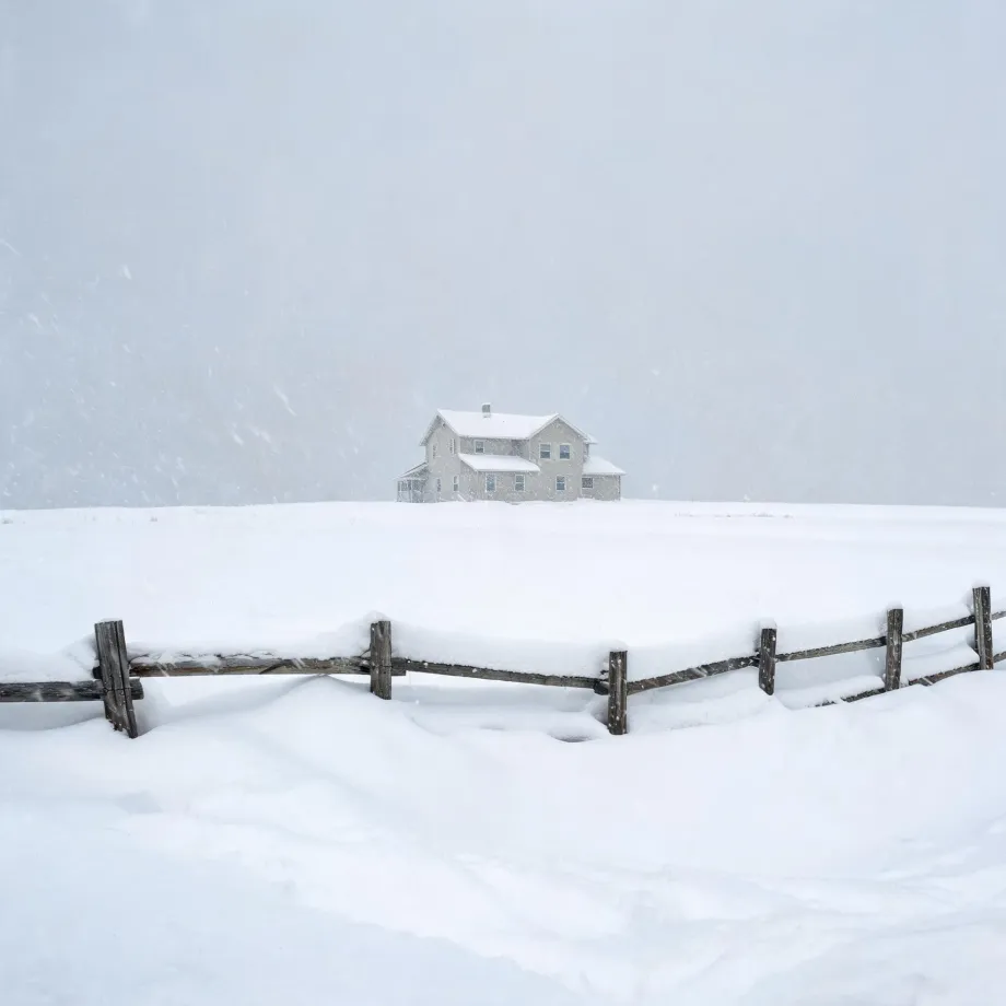 A two-story light-colored house on a snow-covered rise under falling snow and pale sky, with a weathered wooden post-and-rail fence half-buried in drifts in the foreground.