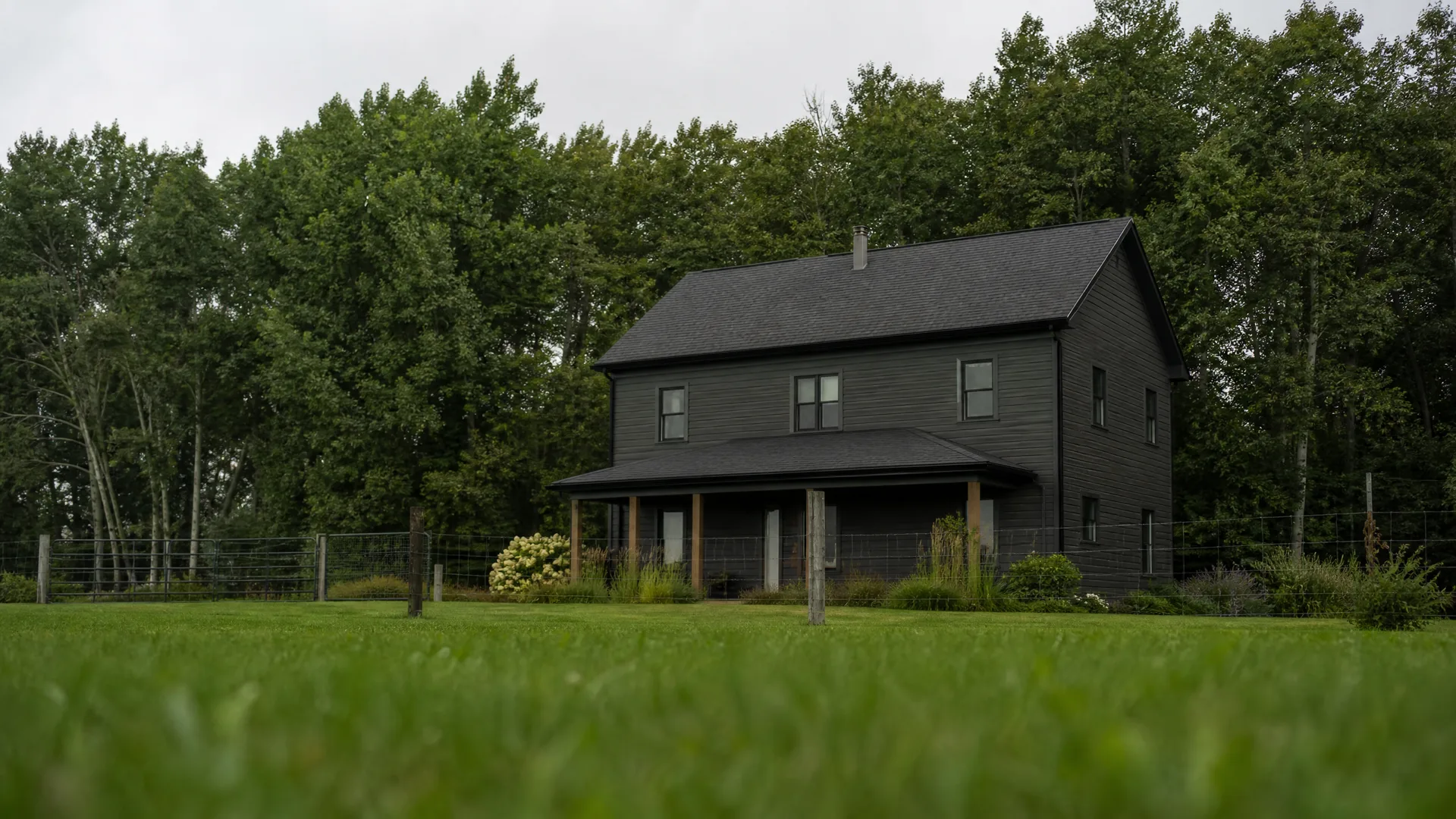 A two-story black-clad house with a gabled roof and covered porch stands on a mown lawn in front of dense green deciduous trees under an overcast sky, with a wire fence and shrubs along the yard.
