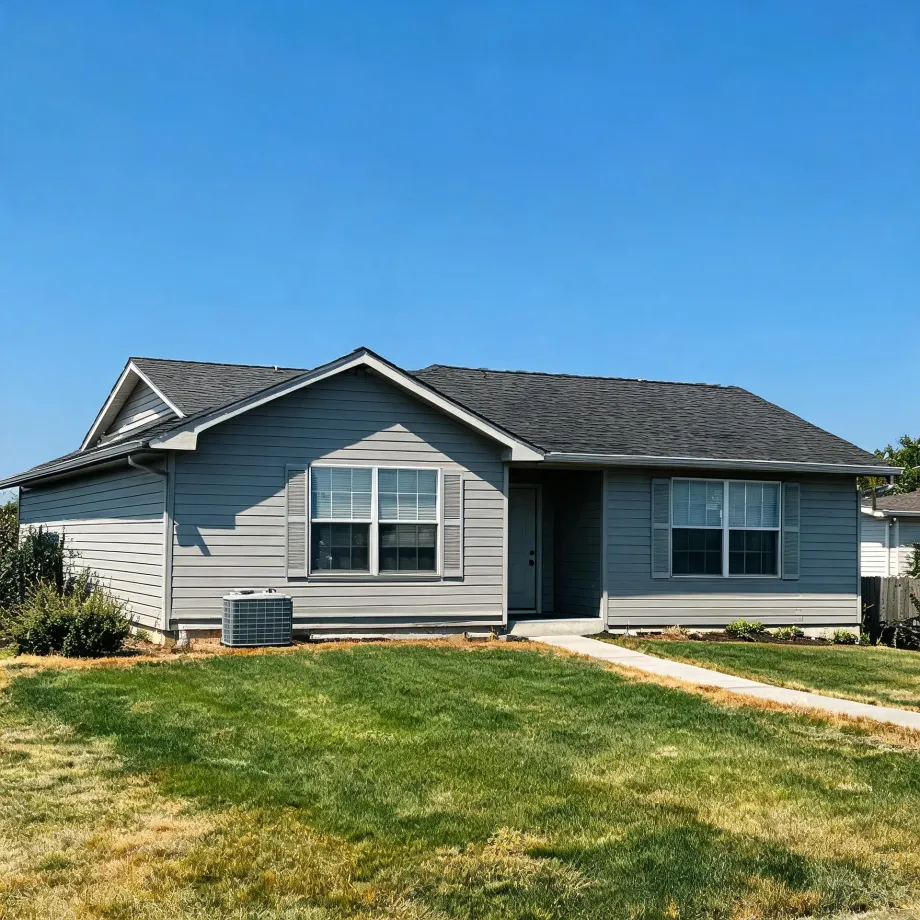 Single-story gray horizontal-siding house with dark shingle roof, white-trimmed windows with shutters, an outdoor air conditioning unit at left, a concrete walkway leading to the front door, and a patchy green and brown lawn under a clear blue sky.