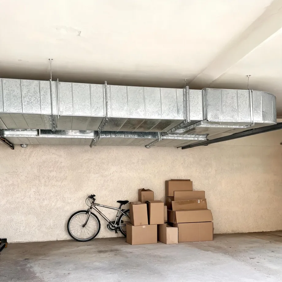 Interior of a garage with galvanized sheet-metal ductwork suspended from the ceiling, a bicycle leaning against a textured beige wall, and a stack of cardboard boxes on the concrete floor.