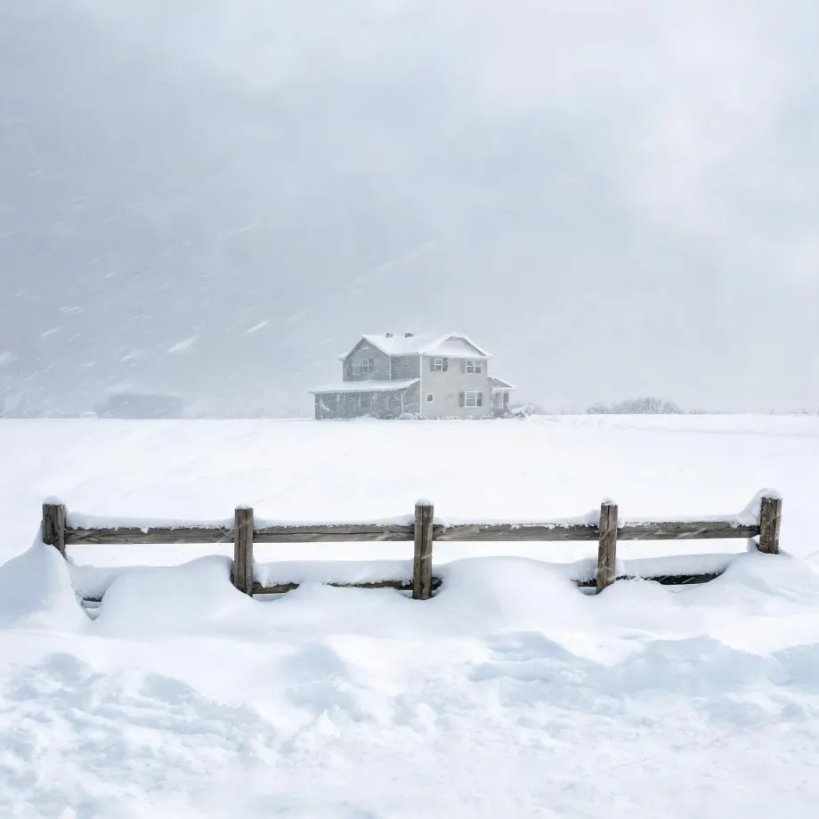 A two-story house with light siding stands in a snow-covered field during falling snow, with a partly buried wooden post-and-rail fence in the foreground and a pale hazy sky behind.