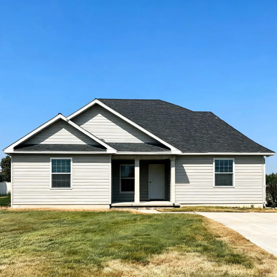 A single-story house with light beige vinyl siding, dark gray gabled shingle roof, a small covered front entry, and two windows, fronted by a patchy green and brown lawn with a concrete driveway at right under a clear blue sky.