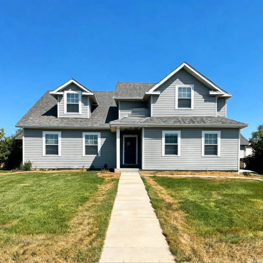 Two-story gray horizontal-siding house with white-trimmed windows, gabled dormers, and a dark front door, fronted by a concrete walkway crossing a partly dry lawn under a clear blue sky.