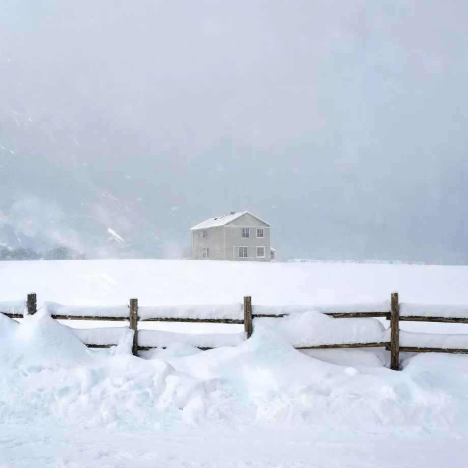 A two-story light-colored house with a snow-covered roof stands on a snowy rise behind a partly buried wooden rail fence during falling snow and low visibility.