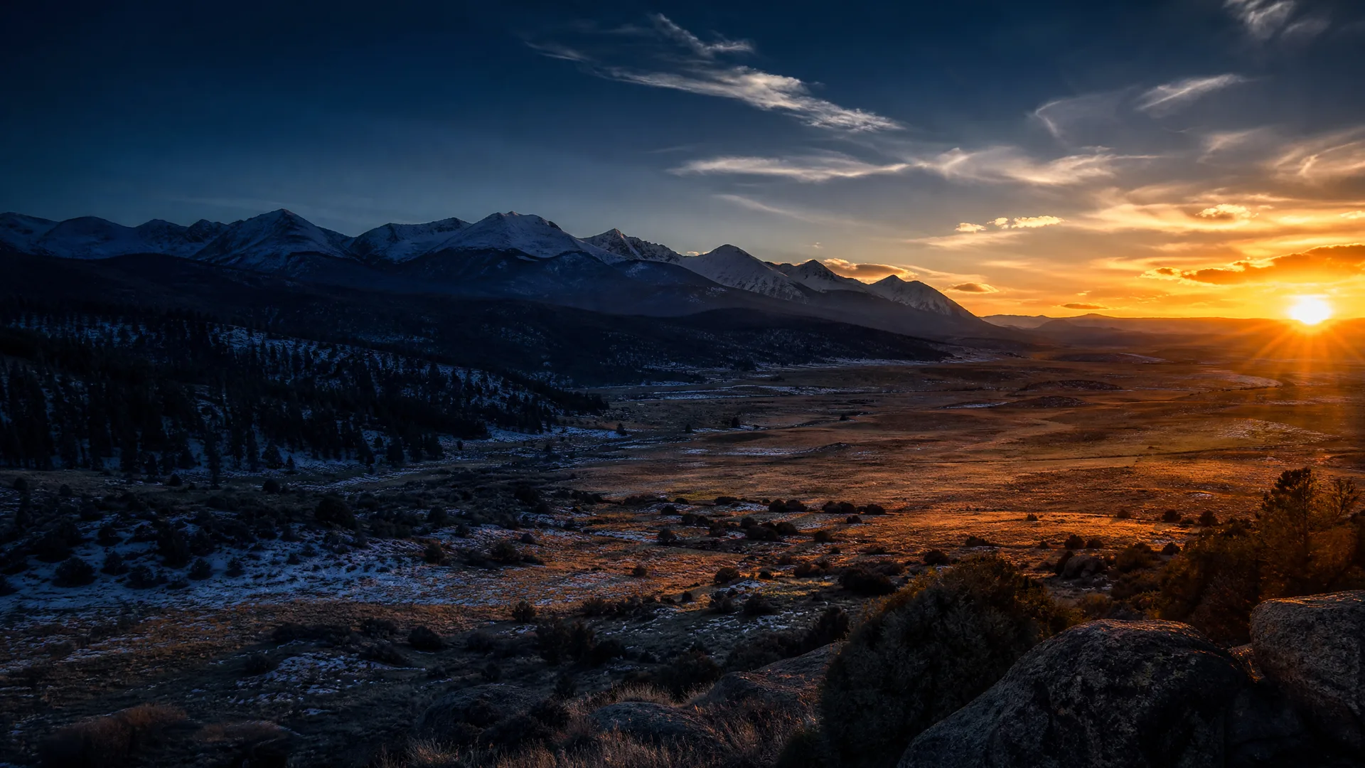 A wide valley at sunset with snow-dusted mountains on the left in shadow, low sun bursting over the horizon on the right, scattered shrubs and rocks in the foreground, and thin clouds in a dark blue sky.