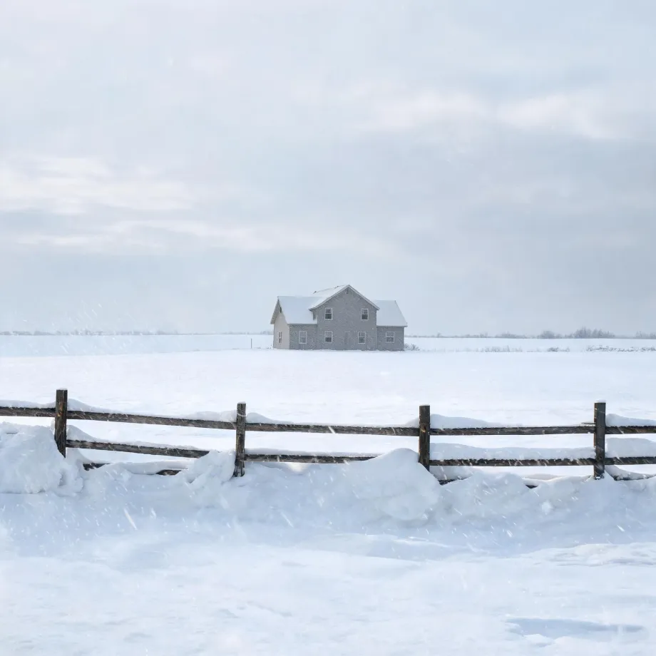 A two-story light-colored house with a gabled roof stands alone in a flat snow-covered field under a pale overcast sky, with a wooden post-and-rail fence and drifted snow in the foreground.