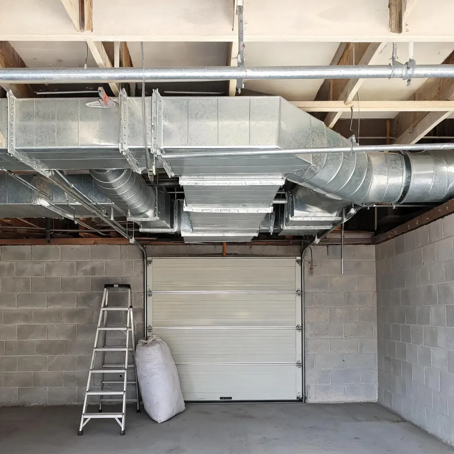 Interior of a garage with concrete block walls and a closed sectional door, showing exposed galvanized sheet-metal ductwork and round ducts mounted under the wood joist ceiling, with a step ladder and a white bag on the floor.