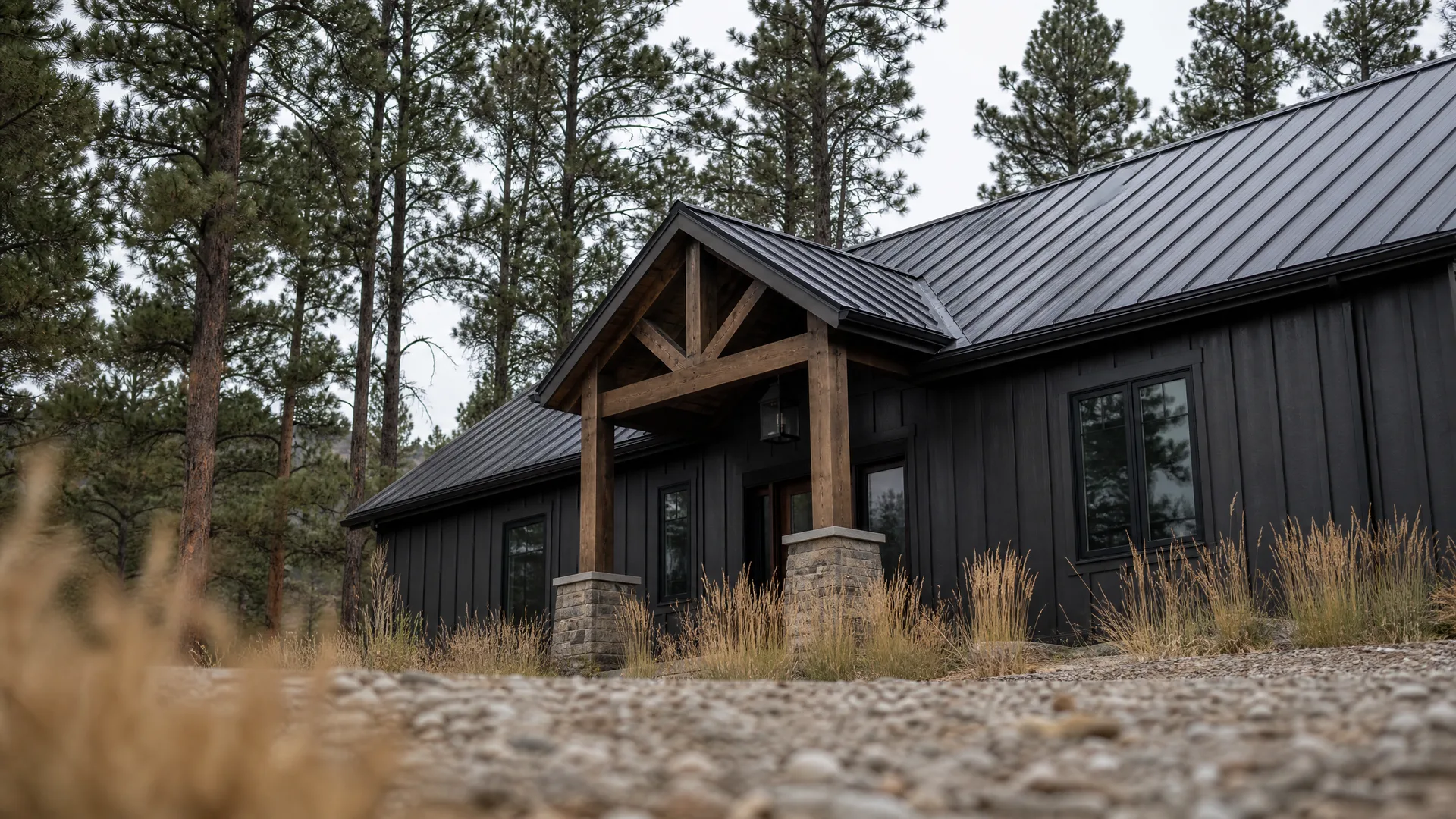 A single-story house with black vertical board-and-batten siding and a dark standing-seam metal roof, featuring a timber-framed entry porch on stone piers, photographed low from a gravel driveway with dry grasses in the foreground and pine trees behind.