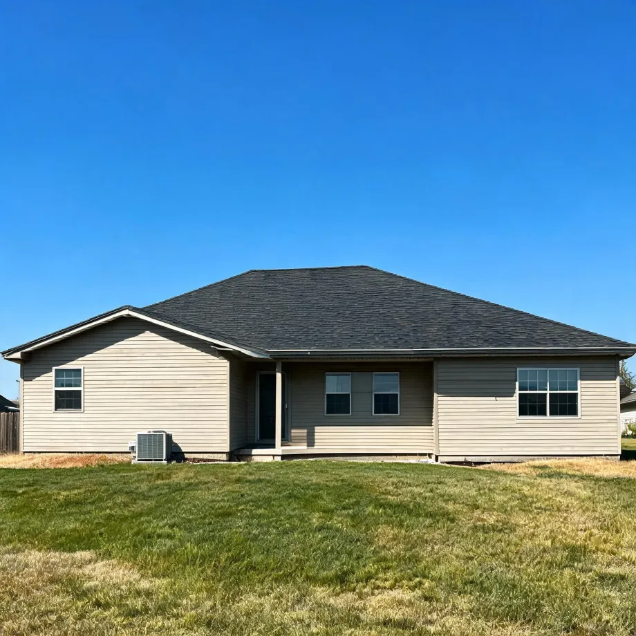Single-story house with tan vinyl siding and a dark shingled hip roof, an exterior air conditioning unit at left, and a mowed grass lawn in front under a clear blue sky.
