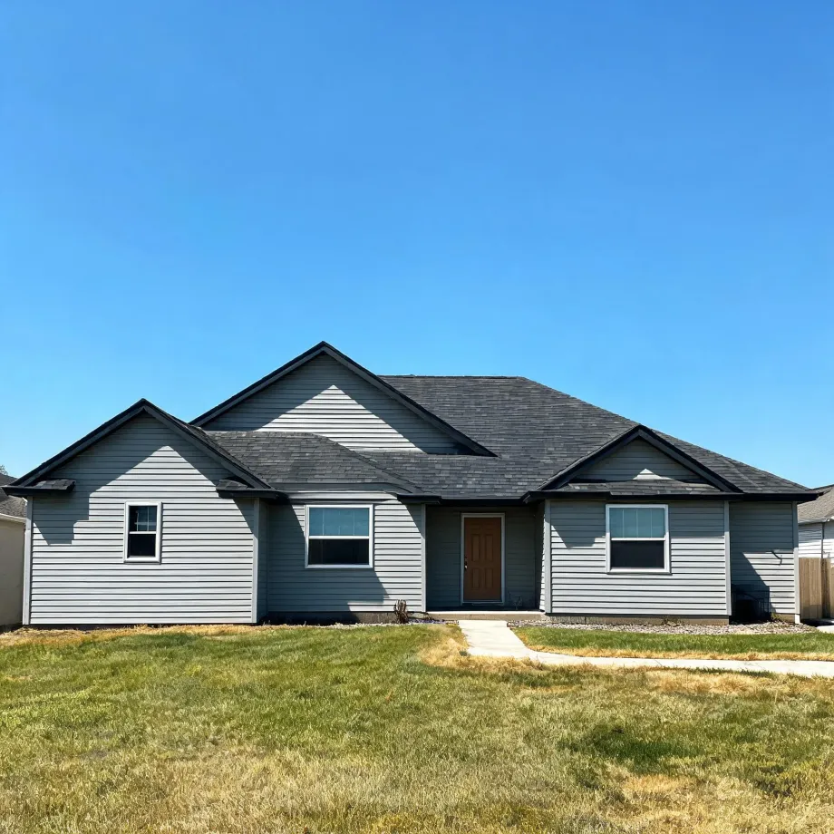 Single-story gray horizontal-siding house with dark shingled gabled roof, a brown front door, and white-trimmed windows, fronted by a patchy green and brown lawn with a concrete walkway under a clear blue sky.