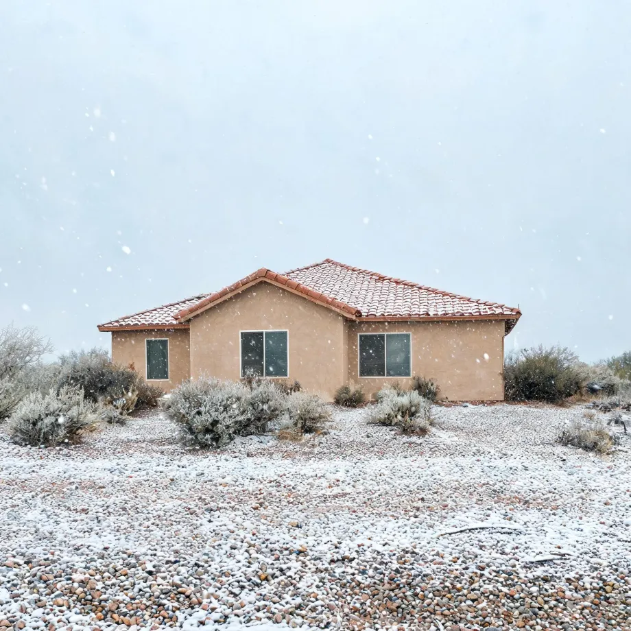 A single-story tan stucco house with a red tile roof during falling snow, surrounded by low desert shrubs and a gravel yard dusted with snow.