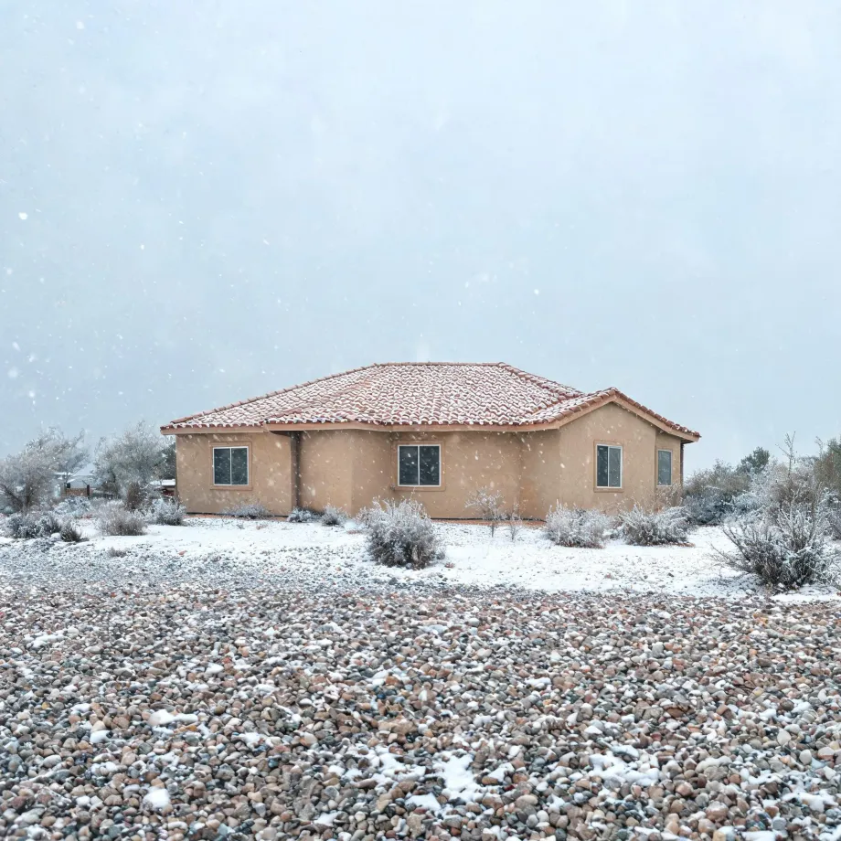 A single-story tan stucco house with a tile roof during falling snow, surrounded by low desert shrubs and a foreground of gravel dusted with snow.