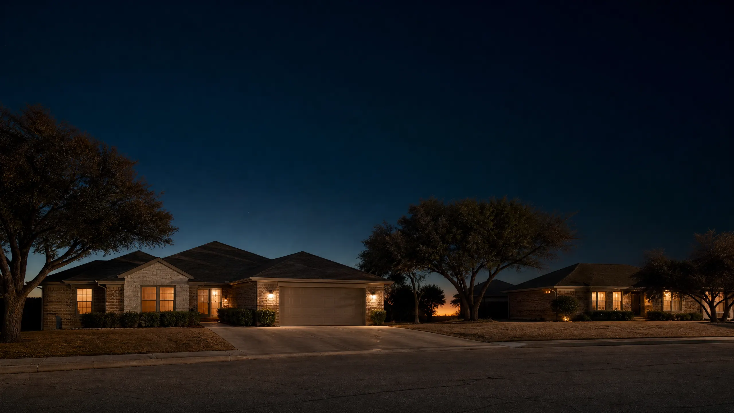 A single-story brick house with a two-car garage photographed at dusk, with interior lights glowing through the windows and trees on either side.