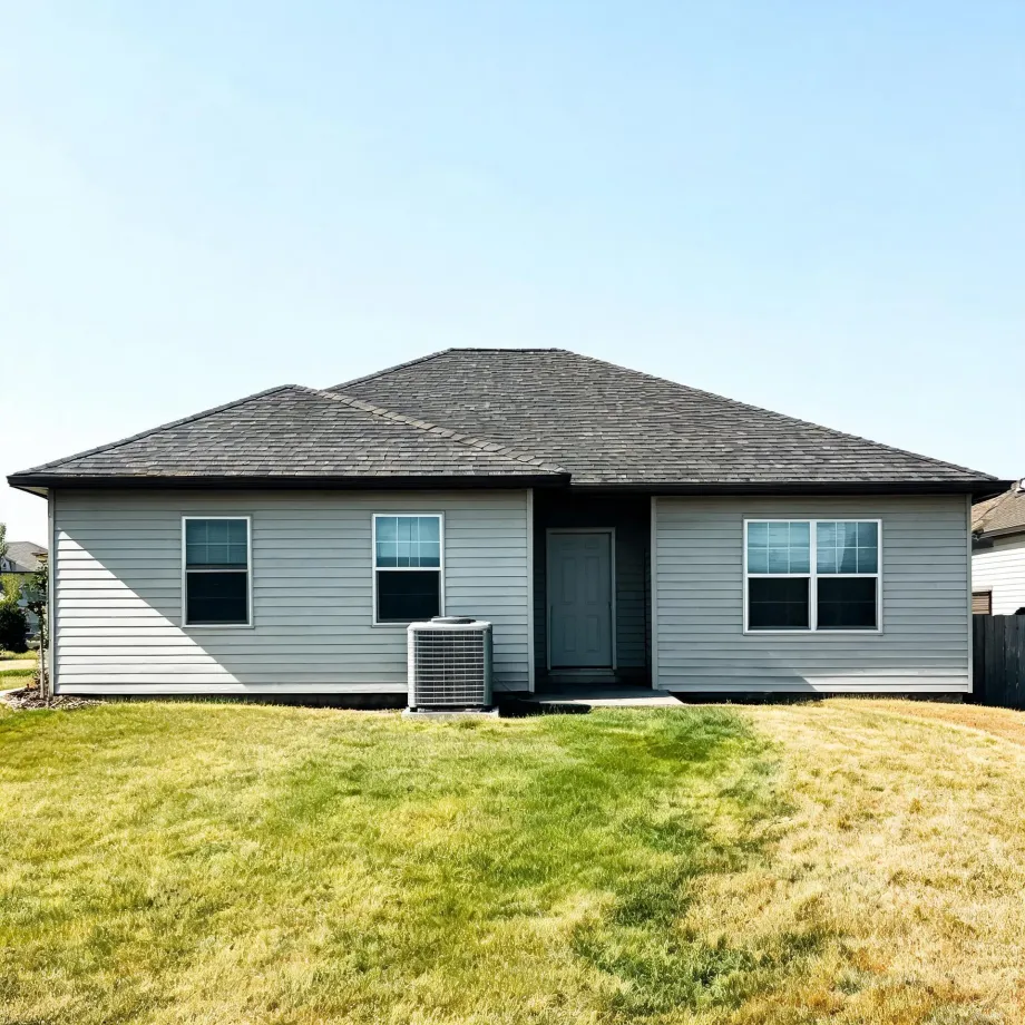 Rear view of a single-story gray vinyl-sided house with a dark shingled hip roof, three windows, a center door, an outdoor air conditioning unit, and a patchy green and brown lawn under a clear sky.