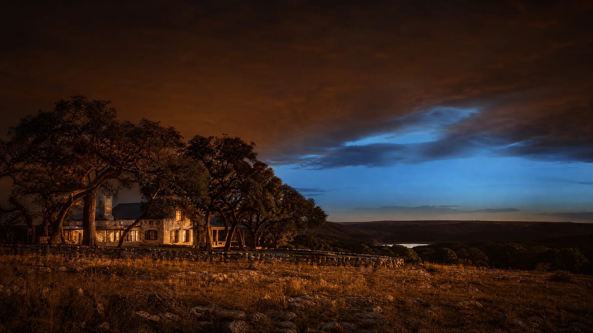 A stone house with lit windows sits among large spreading oak trees on a rocky grass ridge at dusk, with a distant river valley and orange-and-blue clouded sky.