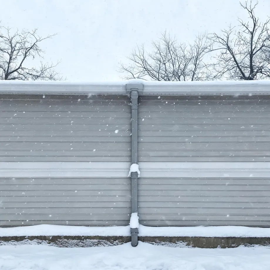A long gray horizontal-siding wall with a central downspout, topped by a snow-covered roof edge, with bare trees behind and snow on the ground during falling snow.