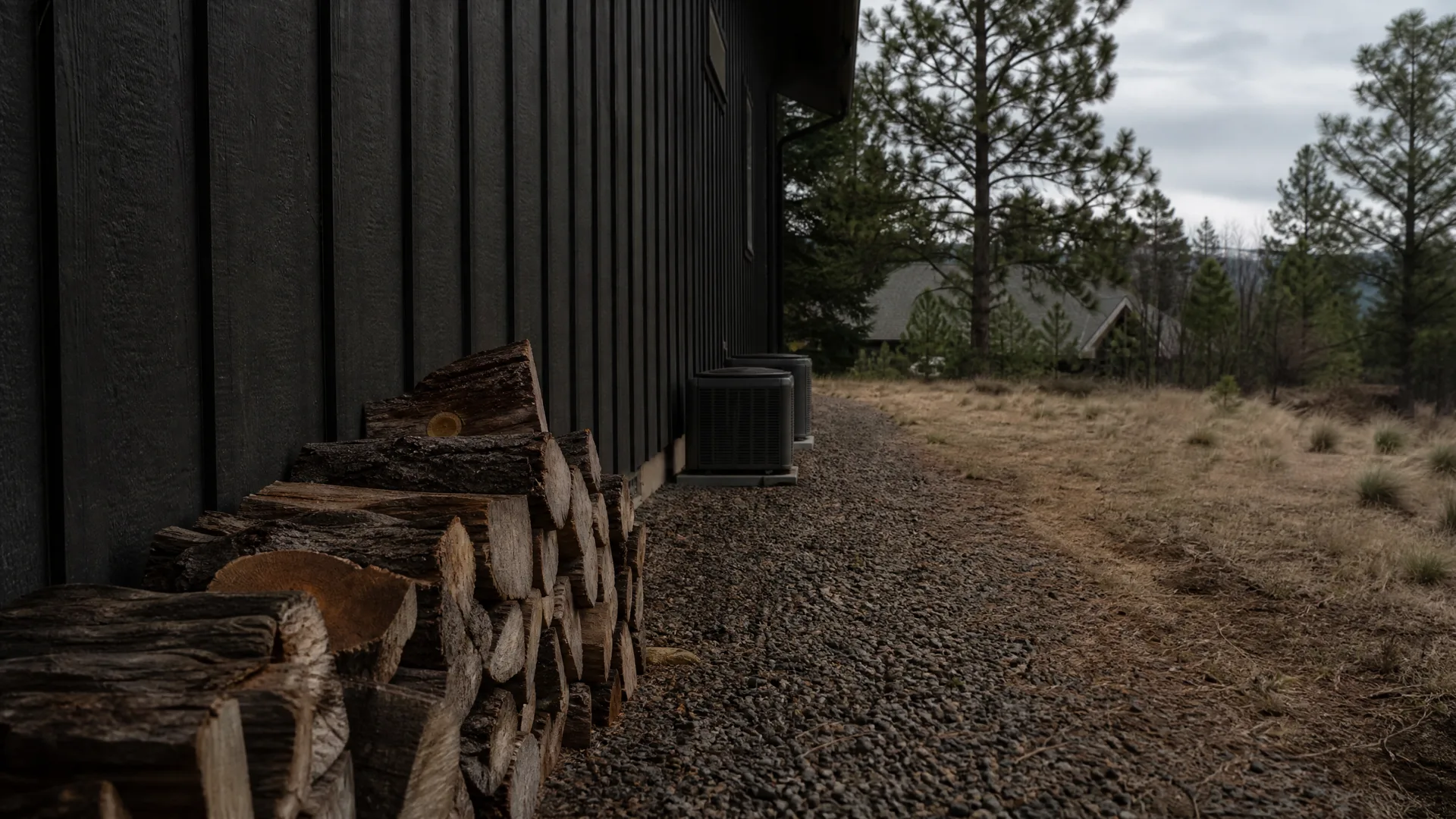 Stacked firewood along the dark vertical-board exterior wall of a house, with two outdoor condenser units, a gravel path, dry grass and pine trees under an overcast sky.