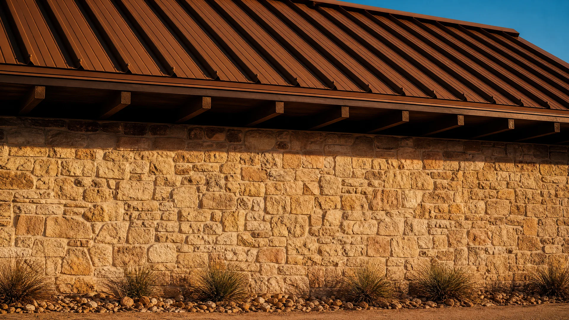 A limestone block wall under a brown standing-seam metal roof with exposed wooden rafter tails, lit by low warm sunlight, with clumps of ornamental grass and a gravel bed at its base.