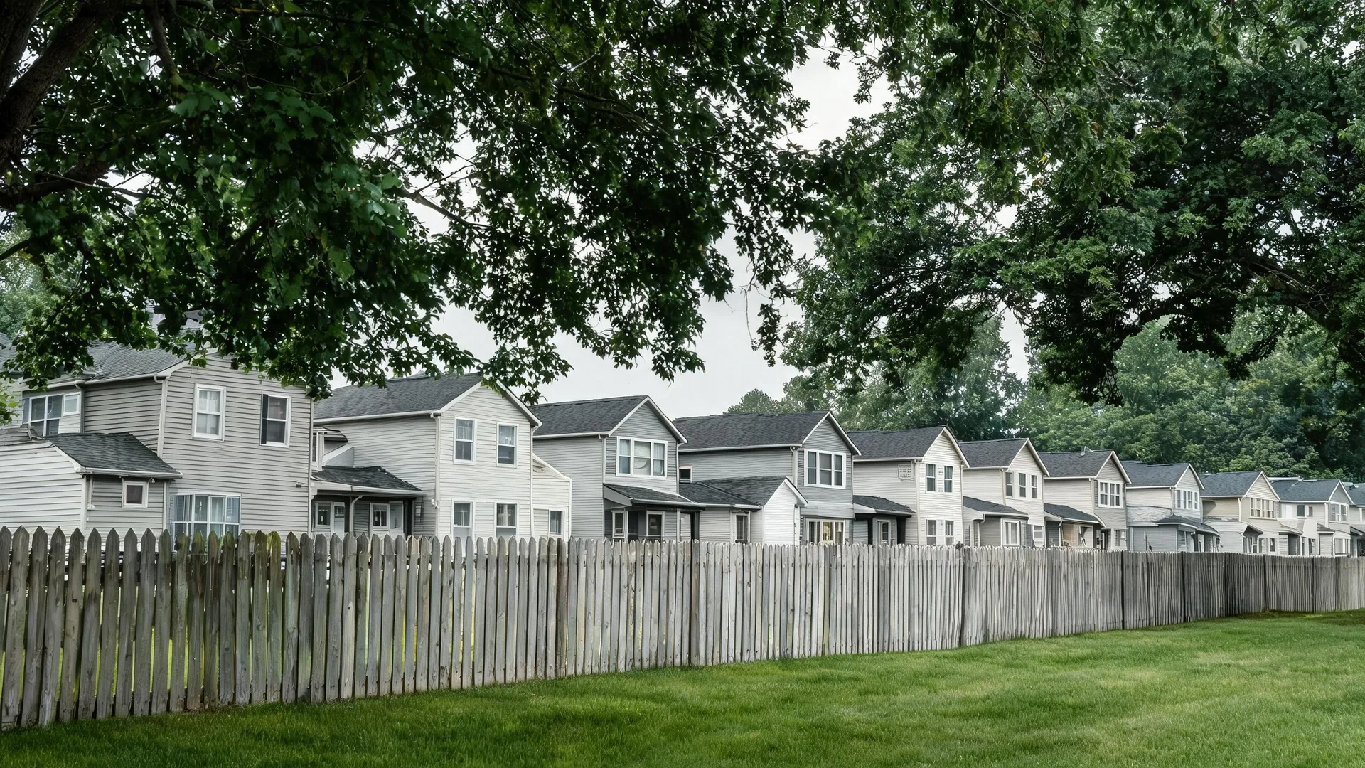 A weathered wooden picket fence runs along a mown lawn in front of a row of two-story light-colored siding townhouses, with large tree branches overhanging under an overcast sky.