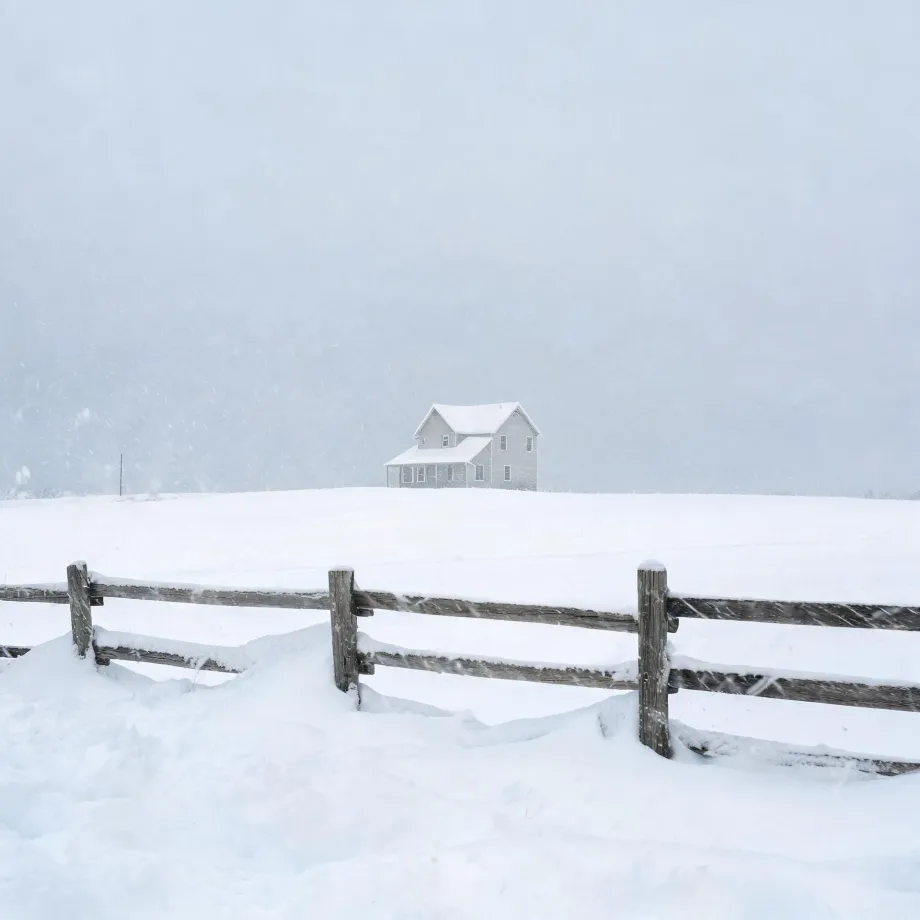 A two-story light-colored house stands alone on a snow-covered rise during falling snow, with a weathered wooden post-and-rail fence crossing the foreground.
