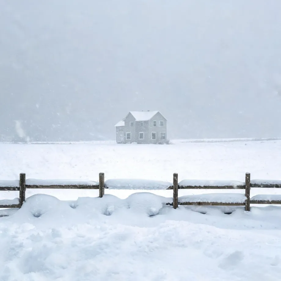 A two-story house with light-colored siding stands alone in a snow-covered field during heavy snowfall, with a snow-topped wooden rail fence crossing the foreground.