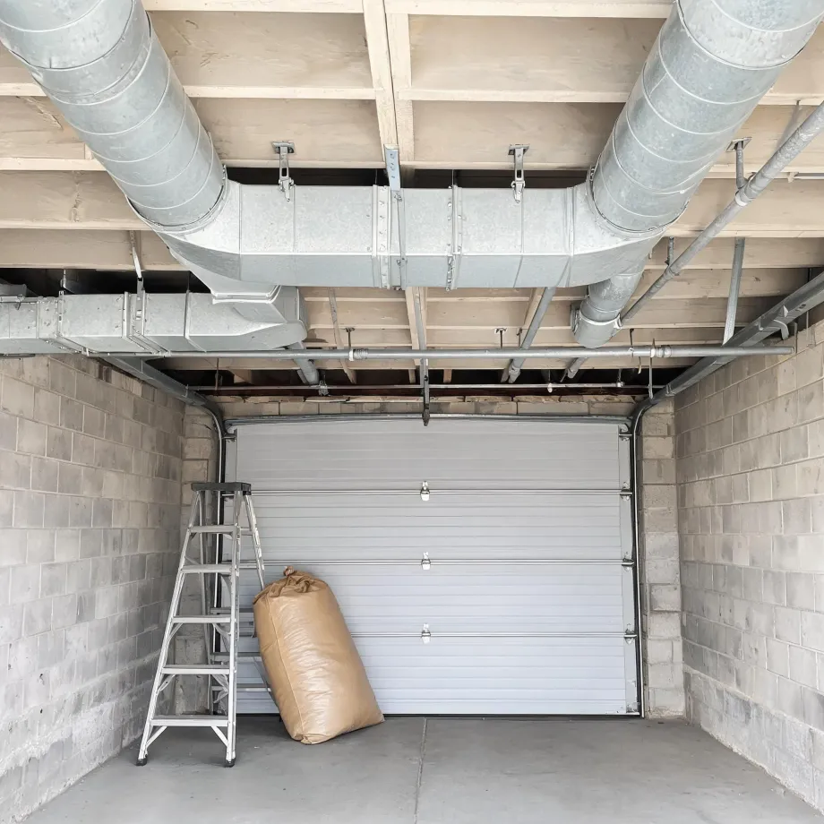 Interior of an empty concrete block garage with a closed white sectional door, an aluminum step ladder and a large tan sack on the floor, and exposed galvanized ductwork and pipes beneath a wood-framed ceiling.
