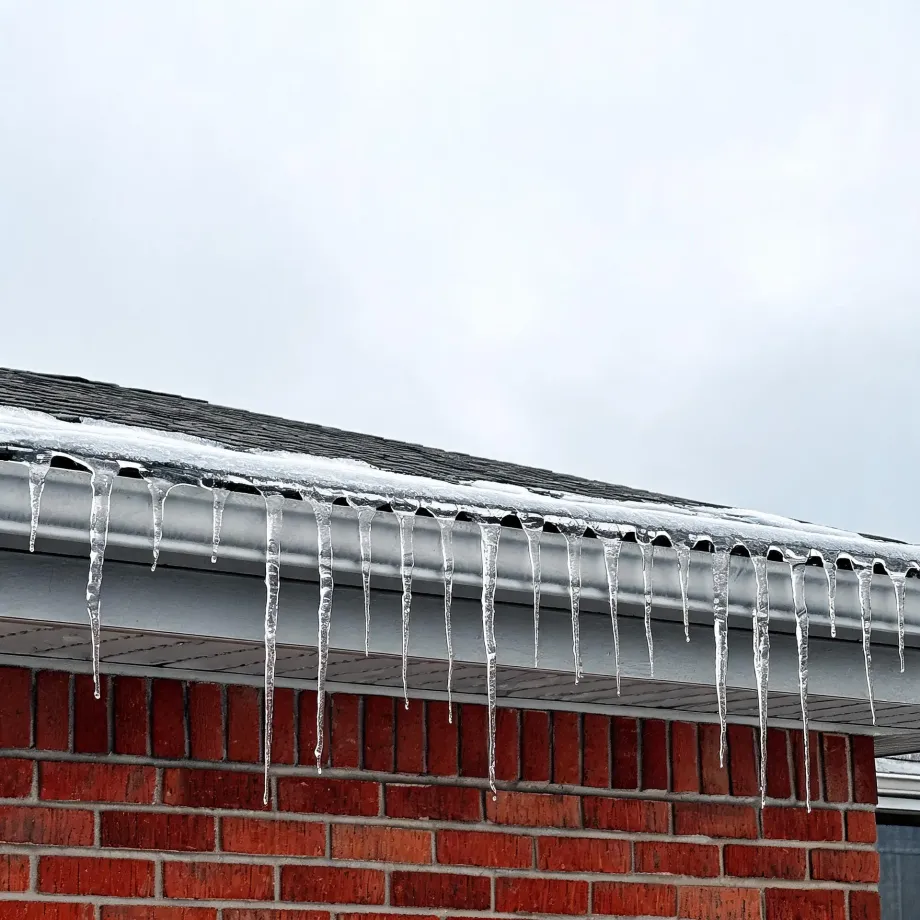 Icicles hanging from a metal gutter along the edge of a shingled roof with snow on it, above a red brick wall, under an overcast white sky.
