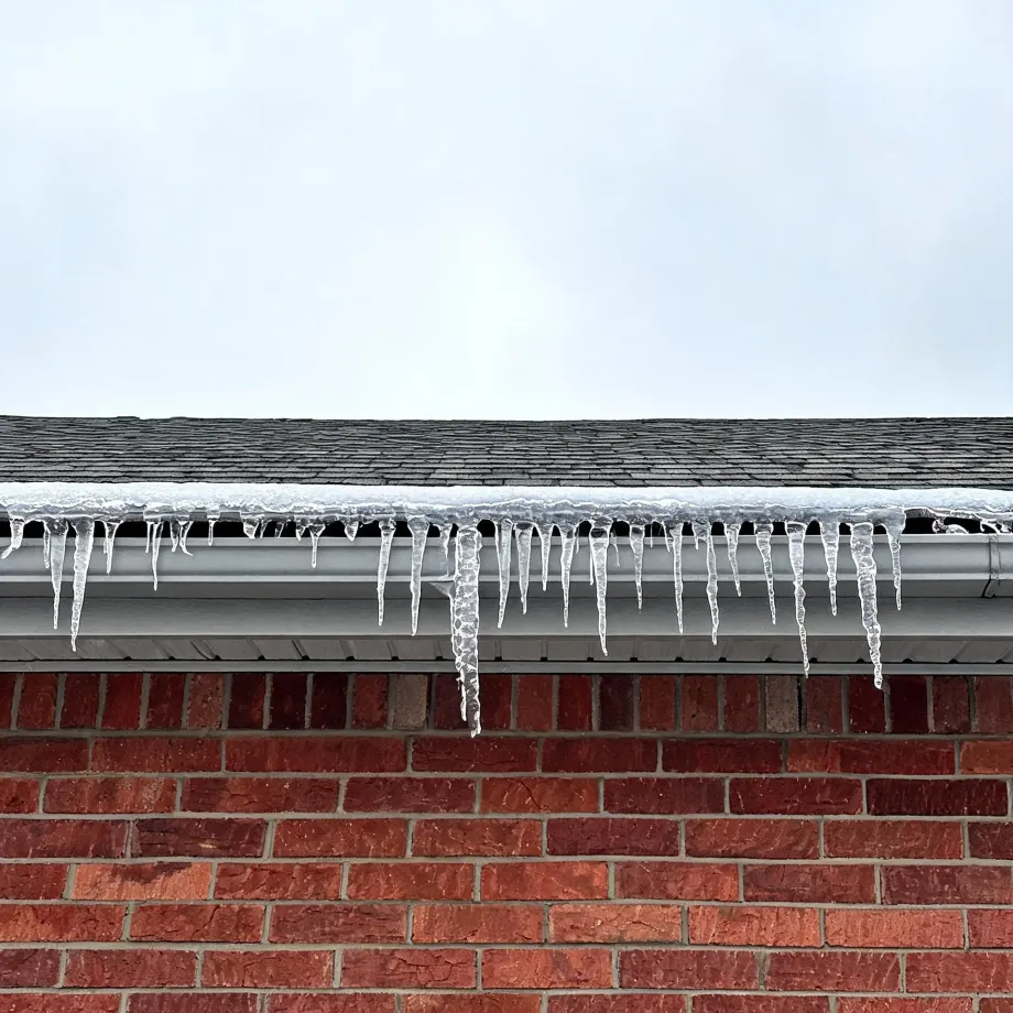 Icicles hang from a white gutter along the edge of a shingled roof above a red brick wall, with an overcast sky behind.