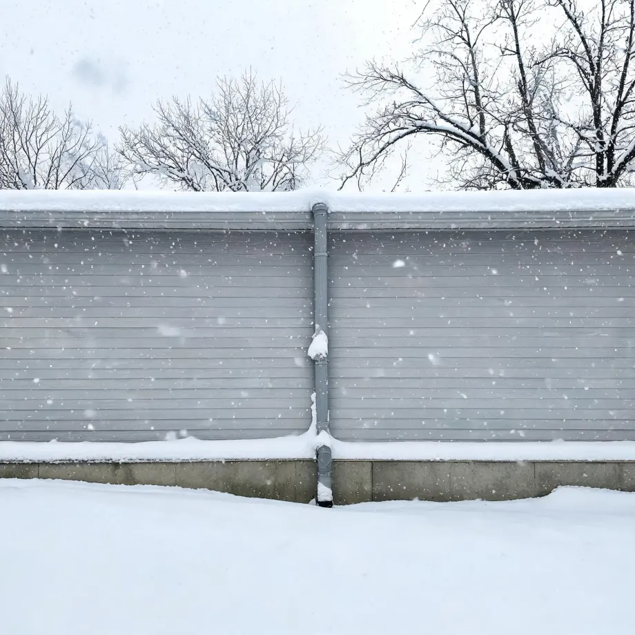 A gray horizontal-sided building wall with a snow-covered roof and a central downspout, seen during falling snow, with bare trees behind and deep snow on the ground in front.