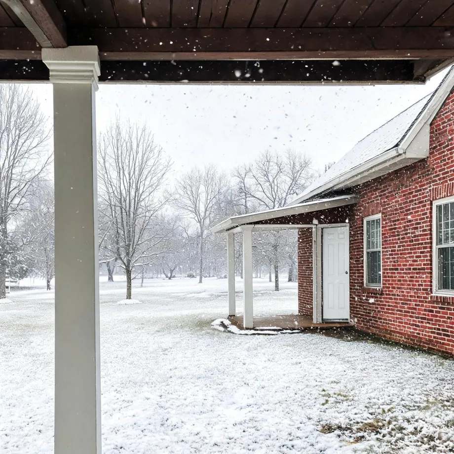 View from under a covered porch toward a red brick house with a white side door and small awning, with falling snow, snow-covered lawn and bare trees in the background.