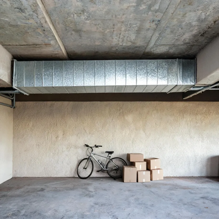 An empty concrete-floored garage or basement with a large galvanized metal duct running below the concrete ceiling, and a bicycle leaning against a textured beige wall beside five stacked cardboard boxes.