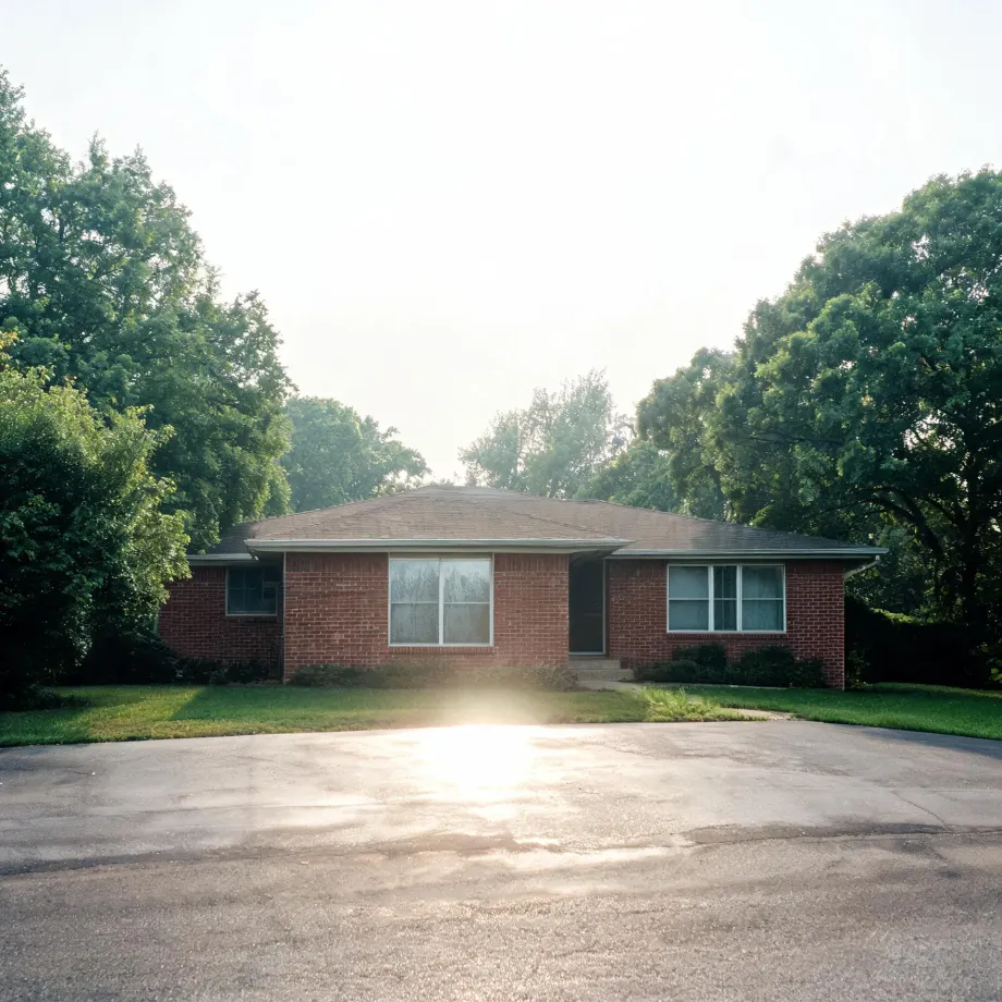 A single-story red brick ranch house with a shingled hip roof, large front window and recessed entry, surrounded by mown grass and mature deciduous trees, fronted by a wide asphalt driveway with sun glare in the foreground.