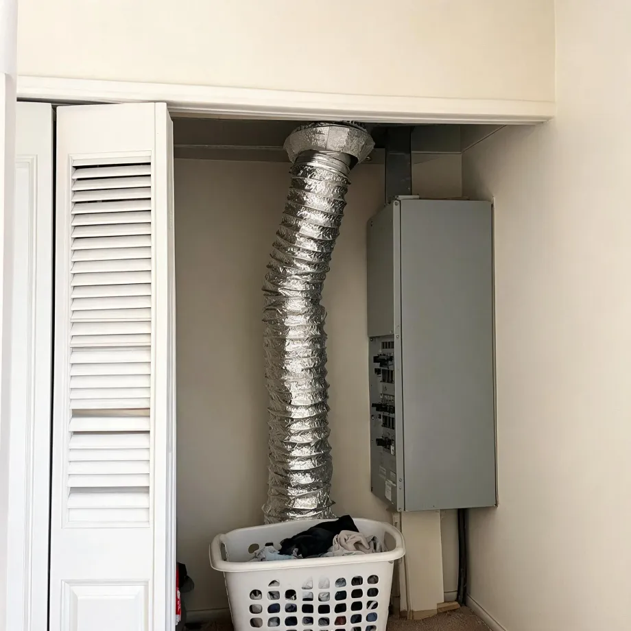 Interior laundry closet with white louvered bi-fold doors, a flexible foil dryer duct running from the floor to the ceiling, a gray electrical panel on the wall, and a white plastic laundry basket of clothes on the floor.