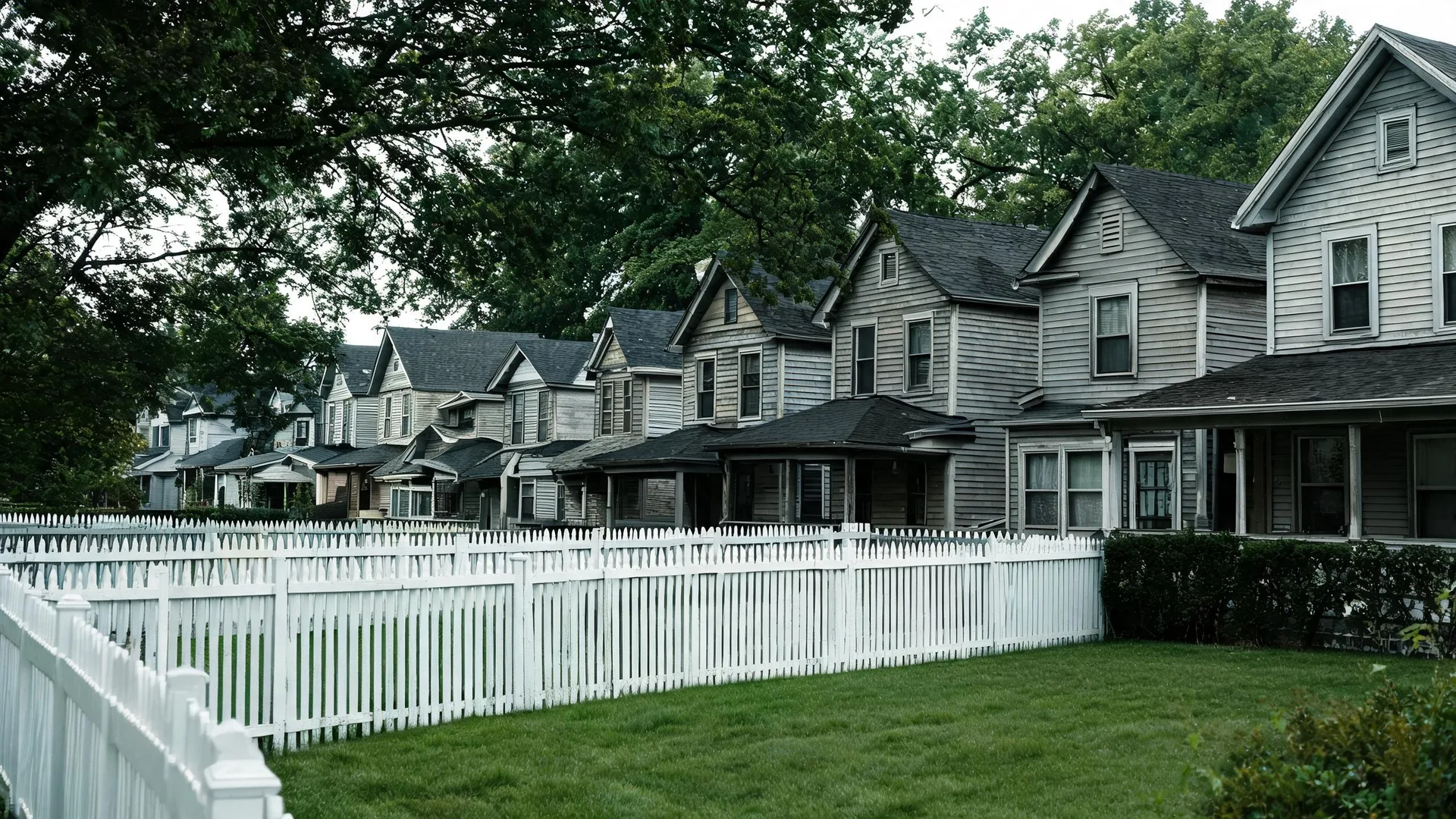 A row of similar two-story wood-sided houses with gabled roofs behind a white picket fence and mown lawn, with large trees overhead.